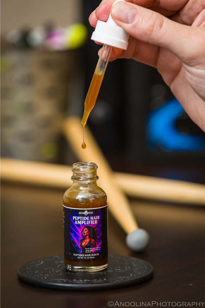 Hand holding a dropper with liquid above a bottle labeled 'Peptide Hair Amplifier' on a blurred background.